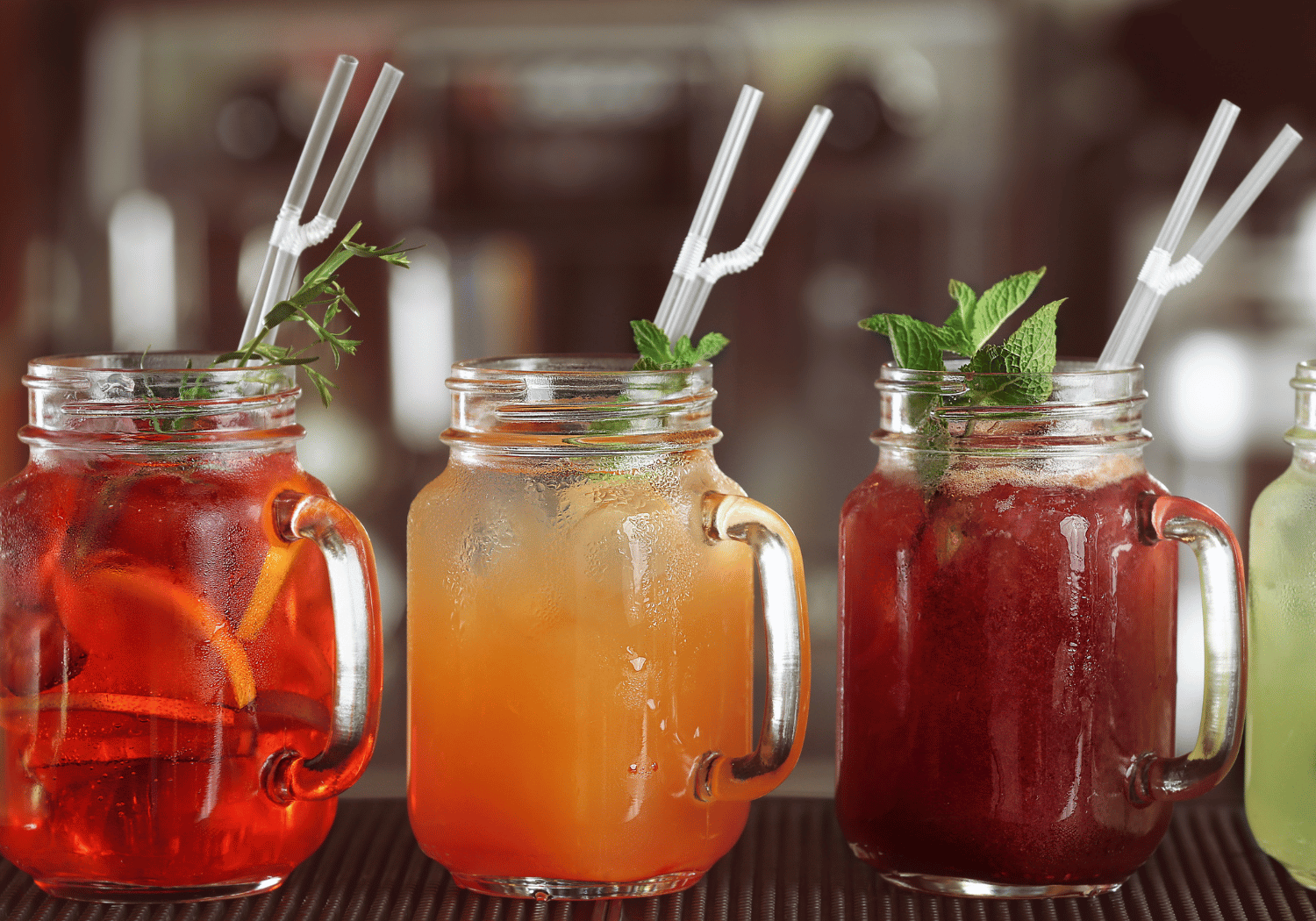 Three colorful iced drinks in mason jars with mint garnish and striped straws on a bar counter